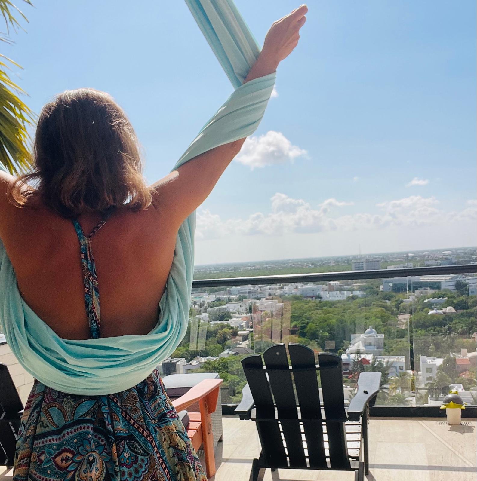 Jenna Jade with her arms raised in pale blue aerial silks on a sunlit balcony.